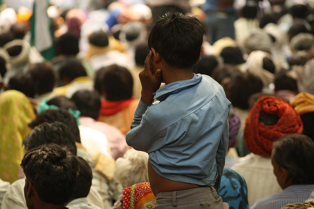 The back of a boy in a blue collared shirt standing up in a crowd of seated people.
