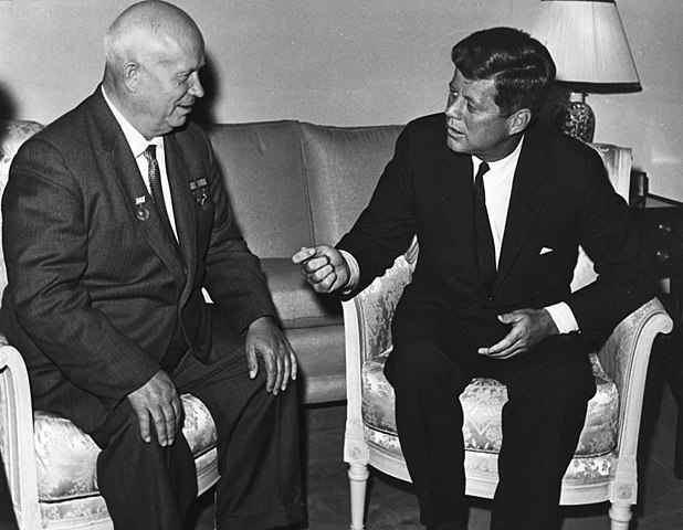 Nikita Krushchev and John F. Kennedy, both wearing suits, sit in armchairs next to each other. Kennedy is speaking and gesturing to Krushchev.