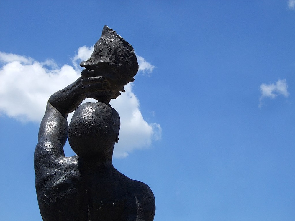A black statue of a man blowing into a conch shell against a blue sky.