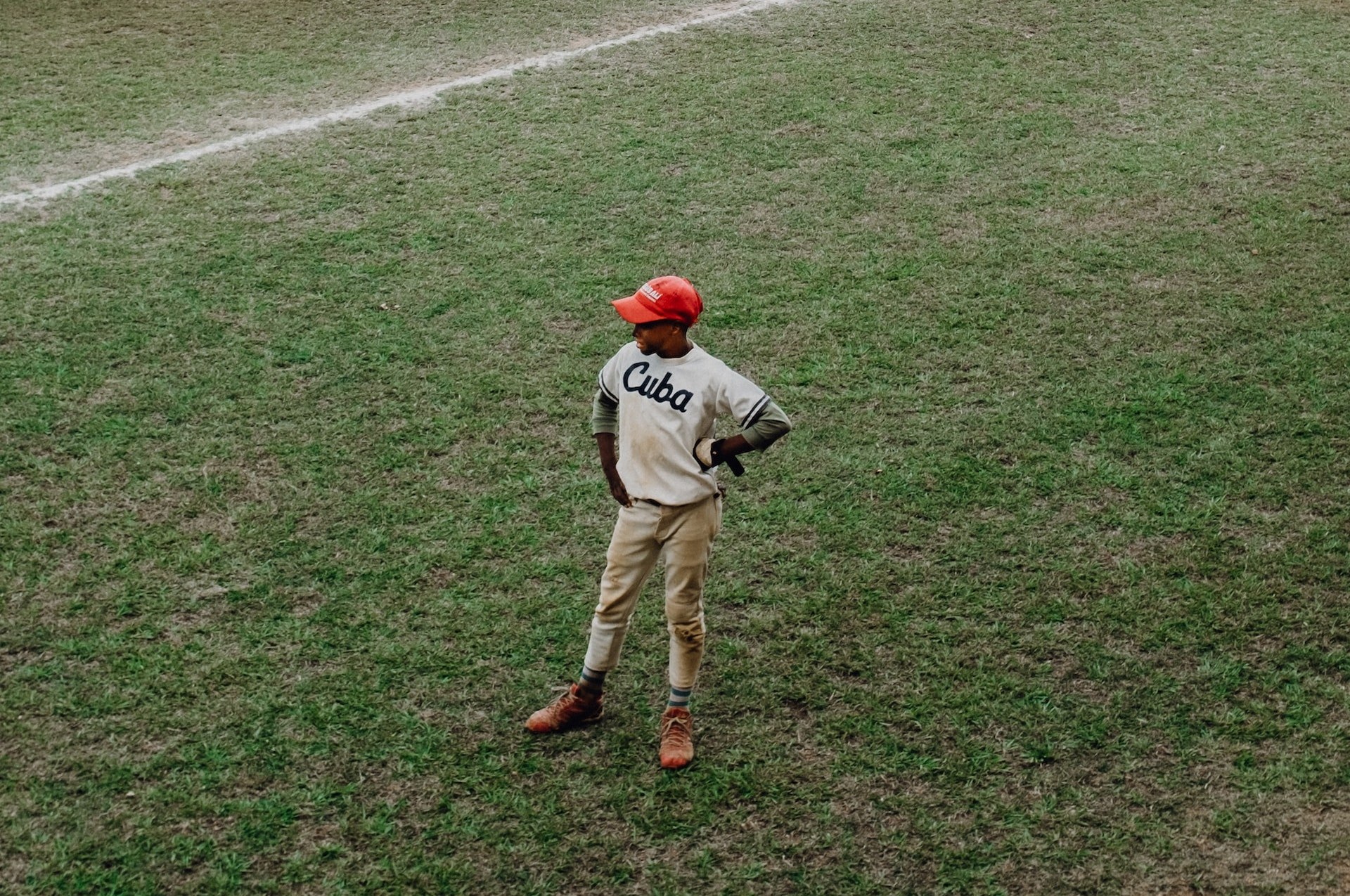 A baseball player wearing a grey "Cuba" uniform and a red baseball cap stands in the outfield with his hand on his hip.