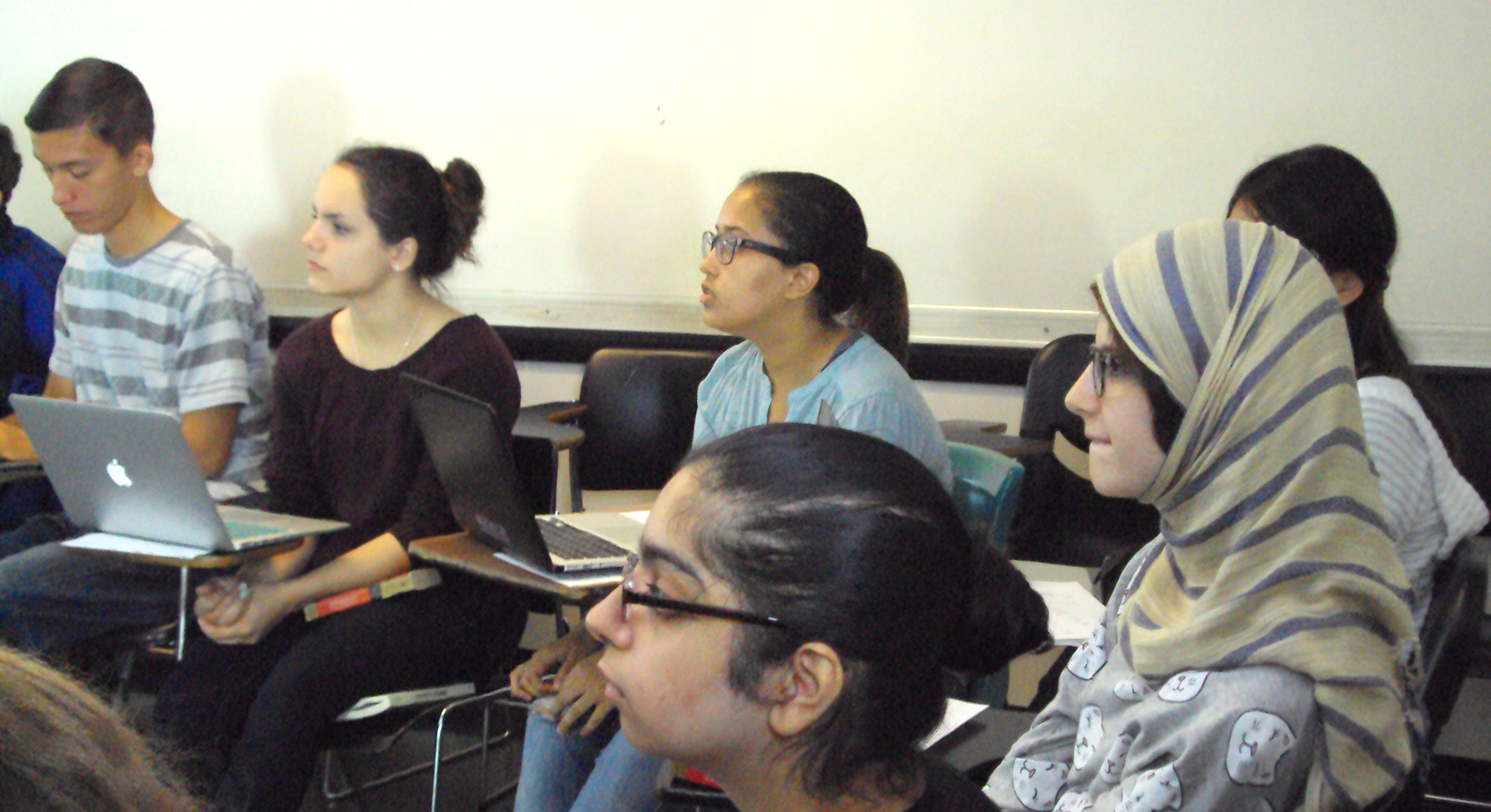 Six students at desks with laptops sit attentively.